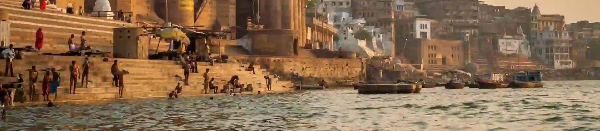 People gathering and bathing along the ghats of the River Ganges in Varanasi during sunset, with historic stone buildings and temples rising along the riverfront