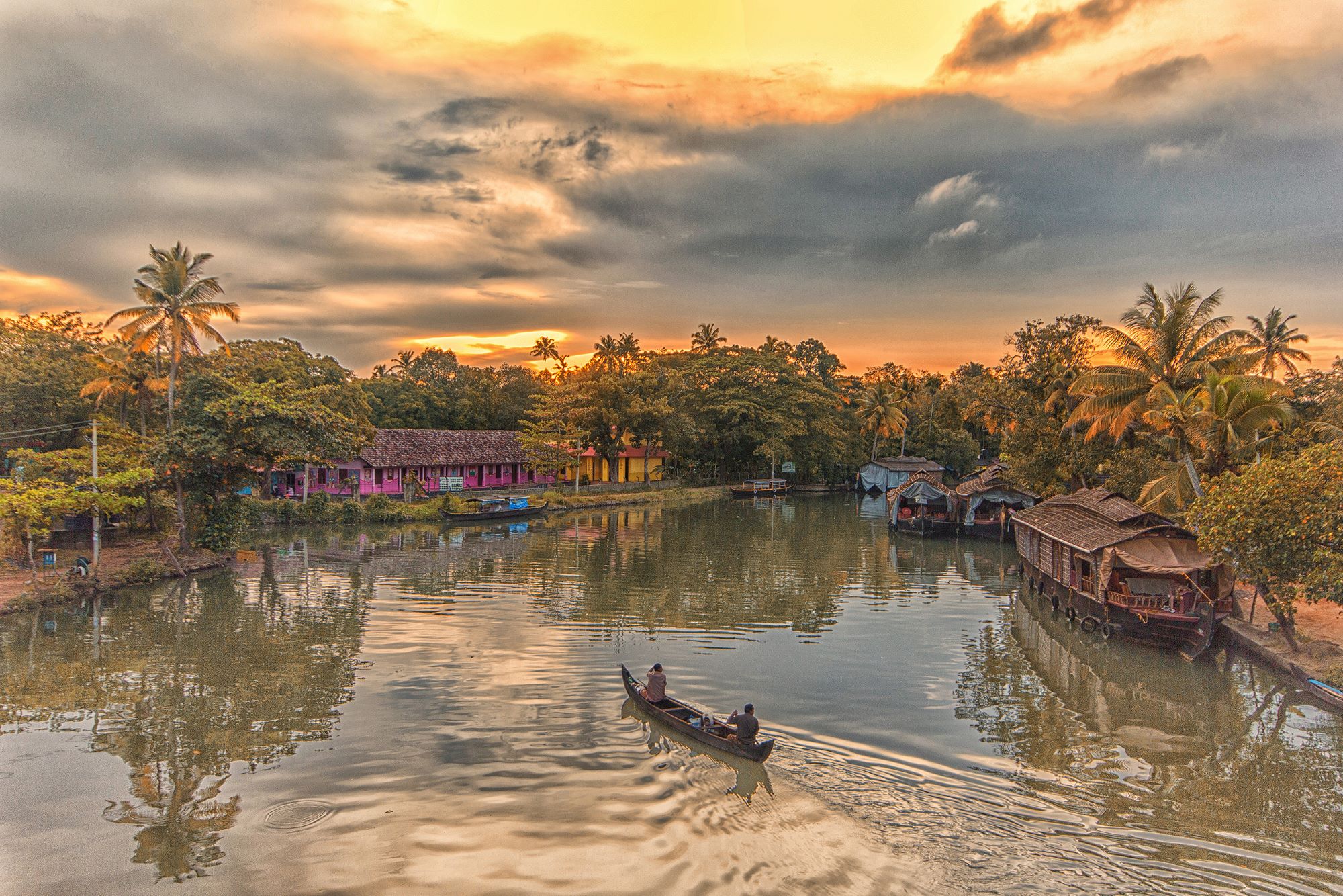 Two people in a canal boat on the Kerala Backwaters at sunset, with a pink house and palm trees lining the banks, and warm light reflecting on the calm water
