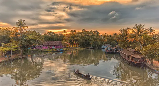 Two people in a canal boat on the Kerala Backwaters at sunset, with a pink house and palm trees lining the banks, and warm light reflecting on the calm water
