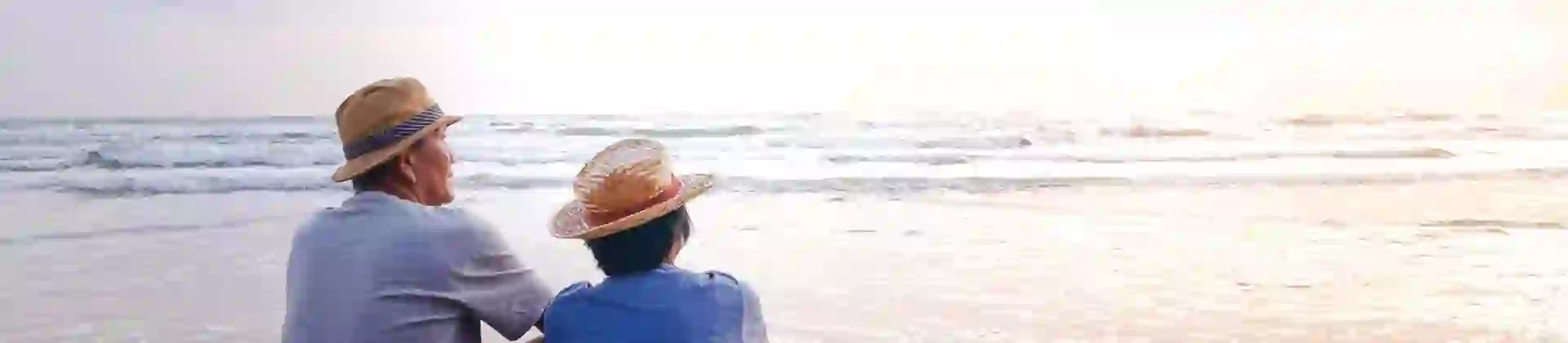 An elderly couple in straw hats sits side by side on a sandy beach, peacefully watching the calm ocean as the sun sets