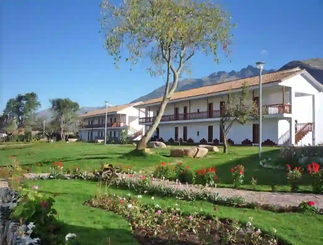 Exterior of Hotel Augustos in Urubamba, featuring a white colonial-style building with gardens, trees, and a mountain backdrop