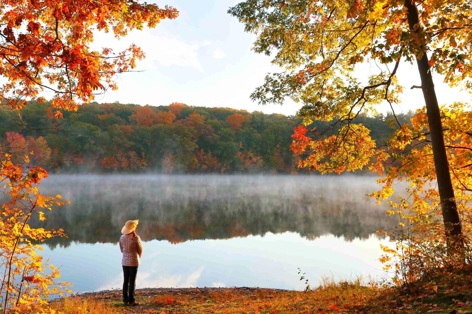 A woman standing by a lake, surrounded by scenic New England autumn foliage with vibrant red, orange, and yellow leaves covering rolling hills