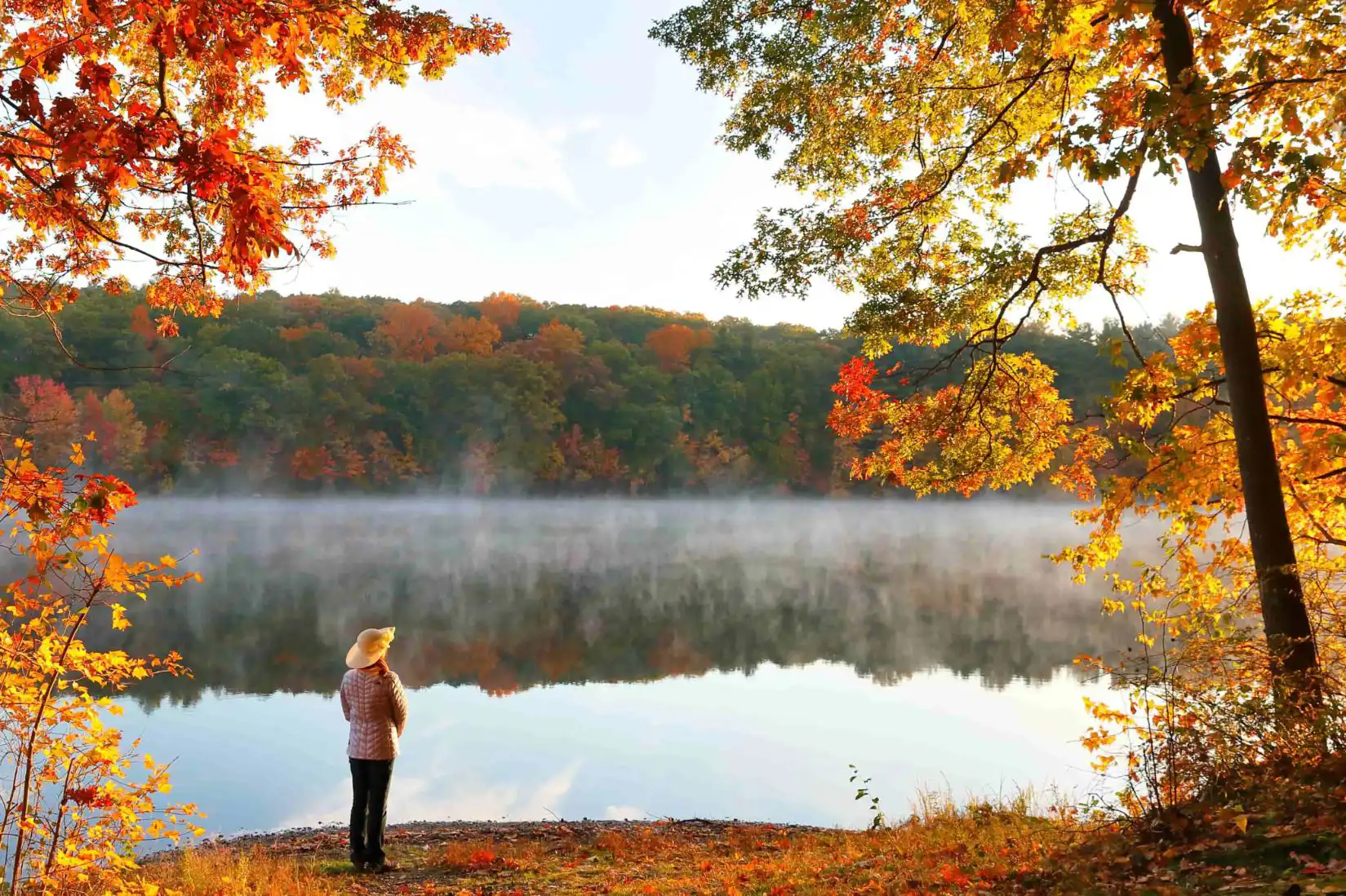 A woman standing by a lake, surrounded by scenic New England autumn foliage with vibrant red, orange, and yellow leaves covering rolling hills