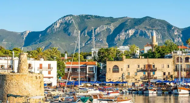 View of a harbour with boats docked, a ruin of a building on the left and buildings further back. Behind, a mountain and a blue sky.