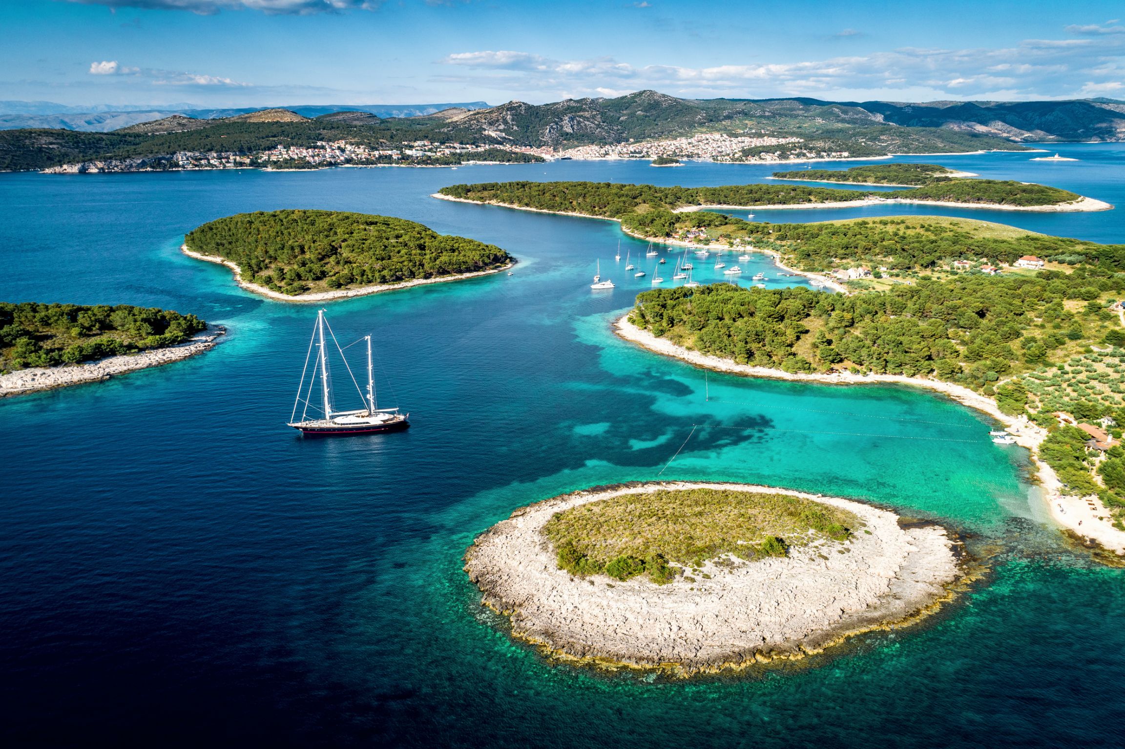 Aerial view of some small islands, surrounded by blue and turquoise water with a sailing boat near the forefront and a few more slightly further away. Mountains in the distance below a blue sky with some clouds.
