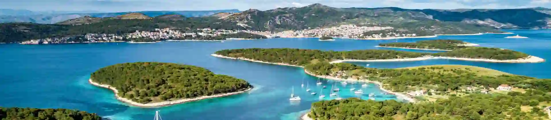 Aerial view of some small islands, surrounded by blue and turquoise water with a sailing boat near the forefront and a few more slightly further away. Mountains in the distance below a blue sky with some clouds.