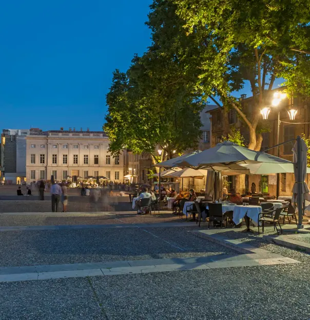 Outdoor seating outside The Palais Des Papes, that has white umbrellas, street lighting and trees above. A cream, wide manor is at the end of the road, and the edge of palace on the left.