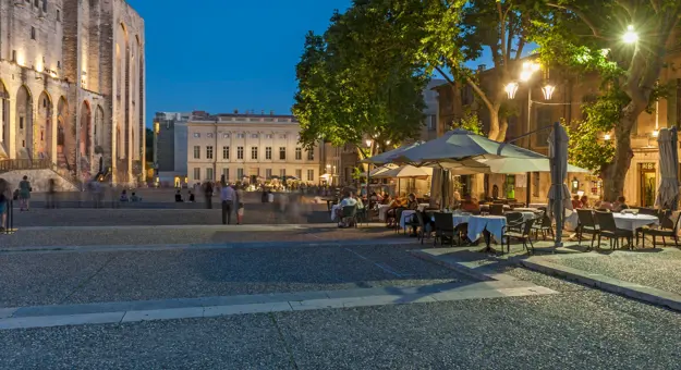 Outdoor seating outside The Palais Des Papes, that has white umbrellas, street lighting and trees above. A cream, wide manor is at the end of the road, and the edge of palace on the left.