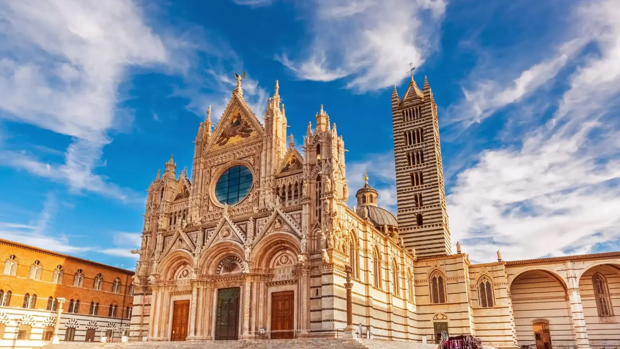 The intricate façade of Siena Cathedral in Italy, bathed in warm sunlight under a vivid blue sky with wispy clouds
