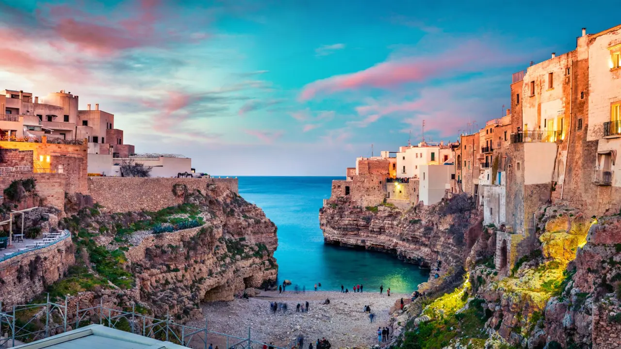 Image looking out to sea with some Polignano's buildings and a beach with people, at sunset