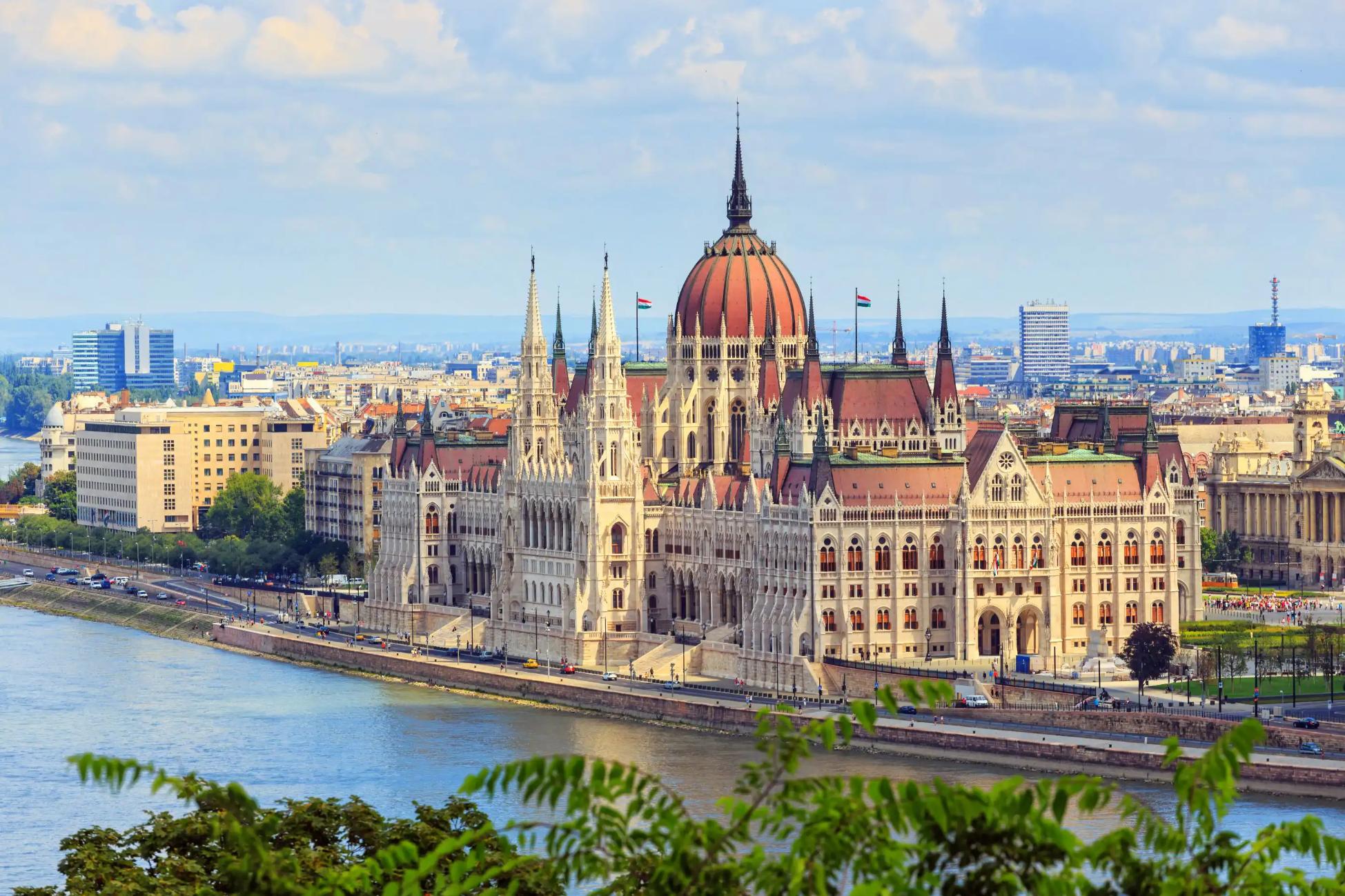 High angle shot of the Hungarian Parliament Building, with red roofs and spiky turrets and the Danube river in the left forefront