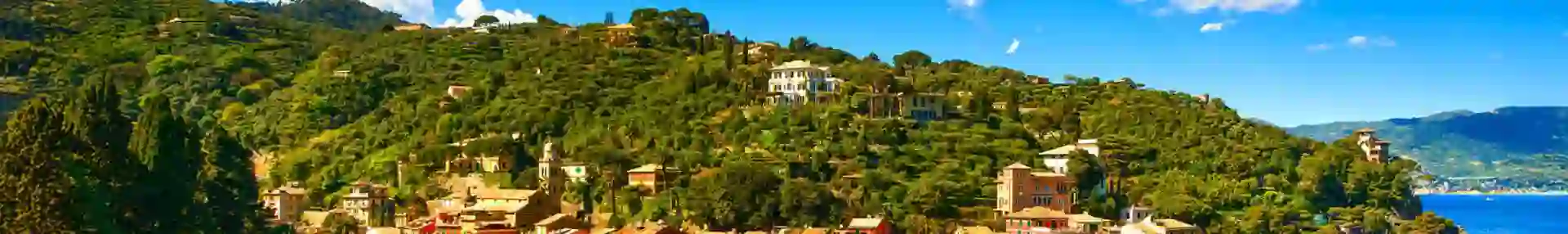 A scenic view of Portofino, Italy, with colourful buildings lining the harbour, boats floating in the clear blue water, and green hills rising in the background under a bright, sunny sky