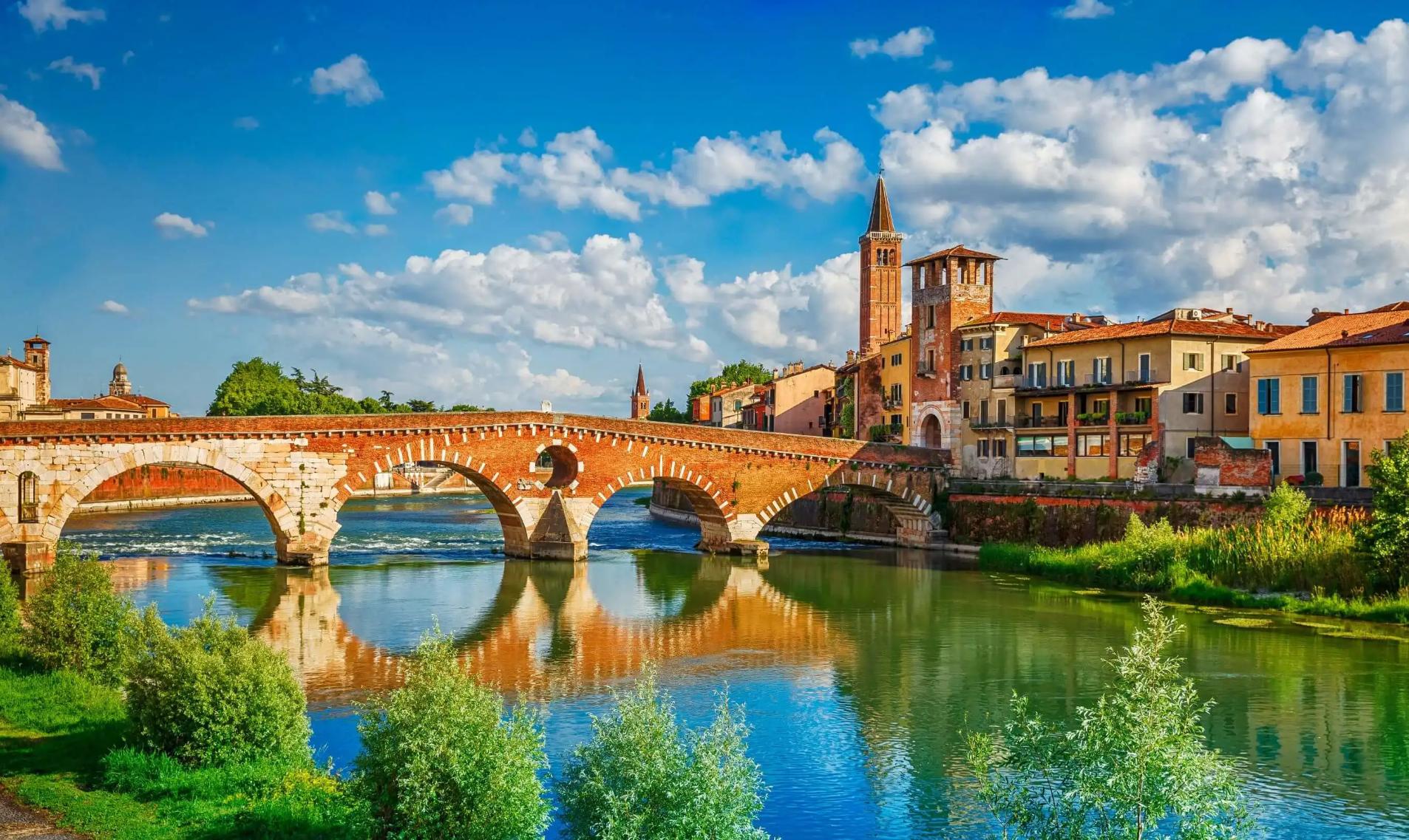 Scenic view of the historic Ponte Pietra bridge crossing the Adige River in Verona, Italy, with colourful old buildings and church towers under a bright blue sky with scattered clouds
