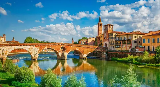 Scenic view of the historic Ponte Pietra bridge crossing the Adige River in Verona, Italy, with colourful old buildings and church towers under a bright blue sky with scattered clouds