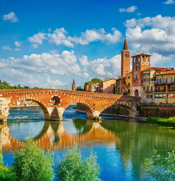 Scenic view of the historic Ponte Pietra bridge crossing the Adige River in Verona, Italy, with colourful old buildings and church towers under a bright blue sky with scattered clouds