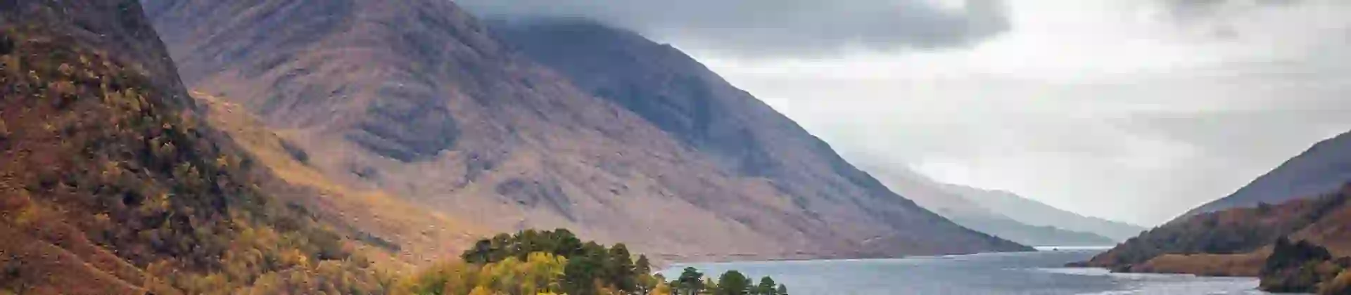 Glenfinnan Monument on a grassy hill at the head of Loch Shiel in the Scottish Highlands, with a lone statue of a Highlander atop the stone tower and dramatic mountains in the background
