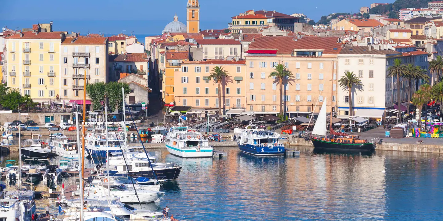View of a harbour from the water, with boats docked to the left and by the land. Beige, cream and yellow buildings on the waterfront with palm trees infront of them. Behind, a slim tower poking out on top of the rest of the buildings, in front of a blue sky.