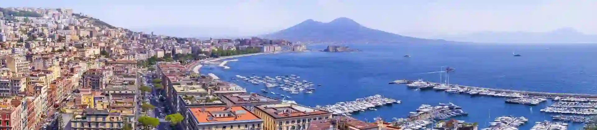 The Bay of Naples with Mount Vesuvius in the distance, lined with waterfront buildings and apartments, and a marina filled with boats