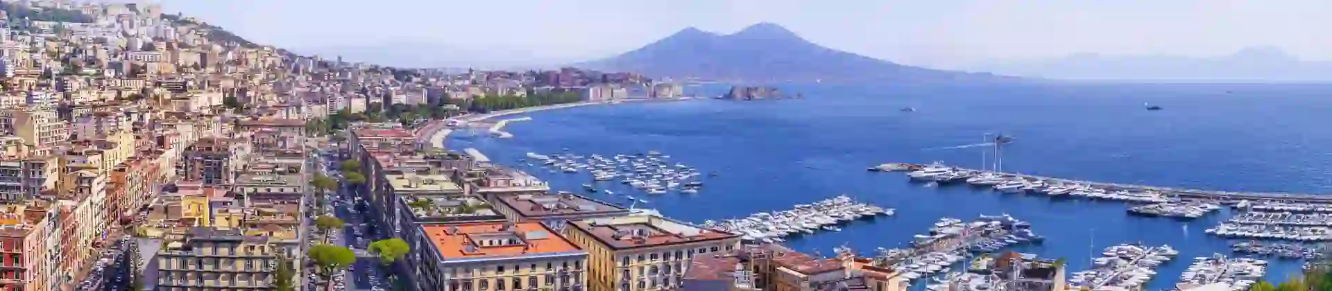 The Bay of Naples with Mount Vesuvius in the distance, lined with waterfront buildings and apartments, and a marina filled with boats