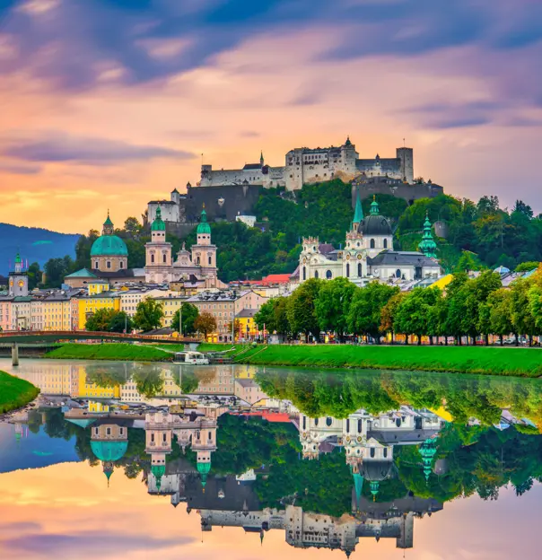 View of Salzburg from the river, showing houses along the river bed, churches and historical landmarks with turquoise turrets behind them, and the fortress on the top of the hill. There is a clear reflection of the city in the river at the forefront of the image, and the sky is a purple and orange sunset