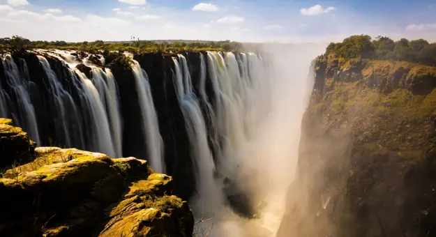 Victoria Falls cascading powerfully over cliffs, surrounded by lush greenery under a bright blue sky with scattered clouds