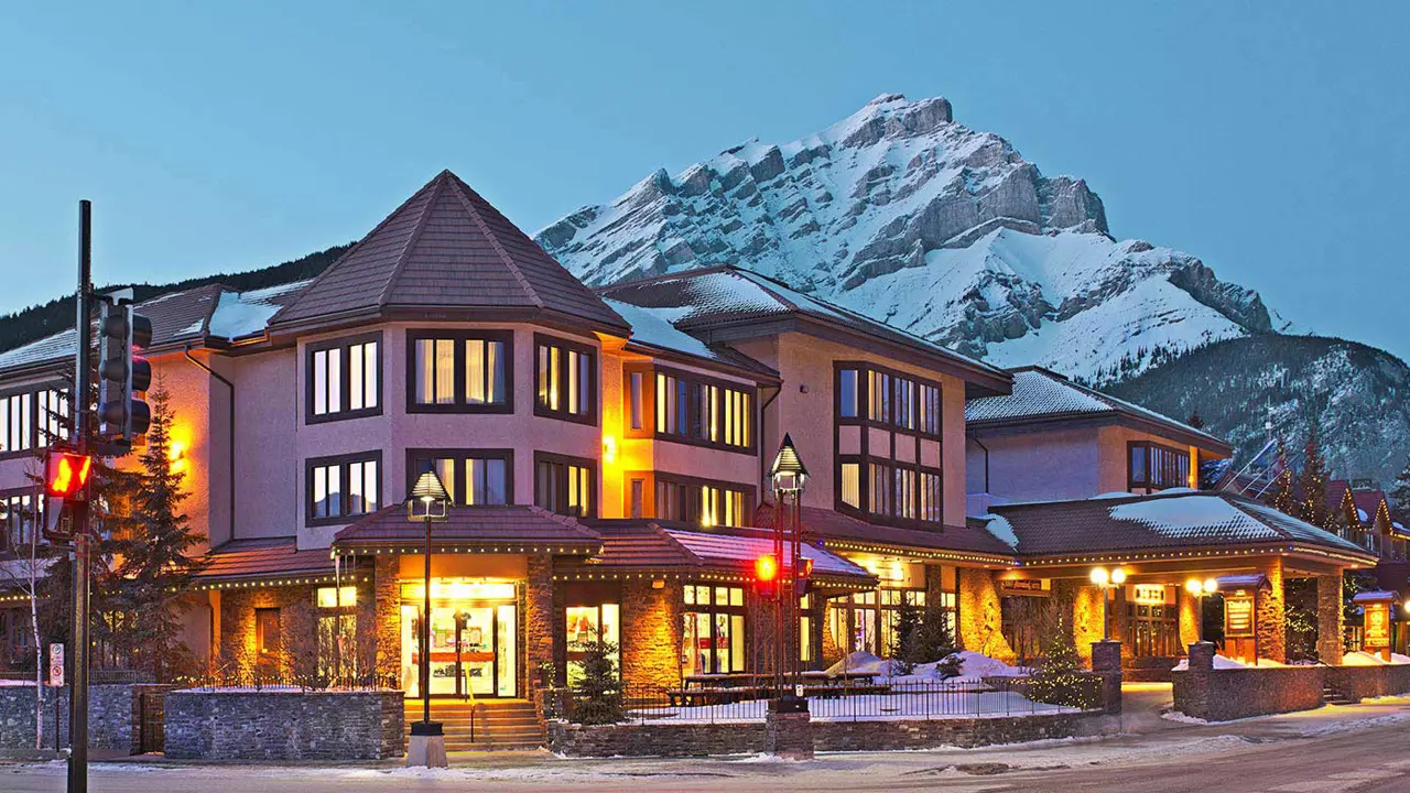 The exterior of Elk + Avenue Hotel in Banff at night, with warmly lit windows and snow-capped mountain in the background
