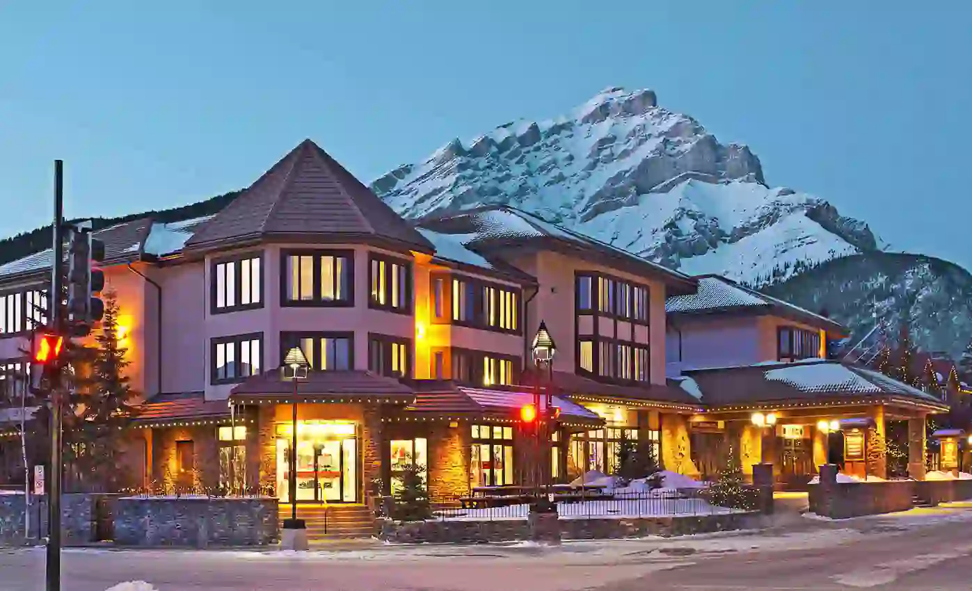The exterior of Elk + Avenue Hotel in Banff at night, with warmly lit windows and snow-capped mountain in the background