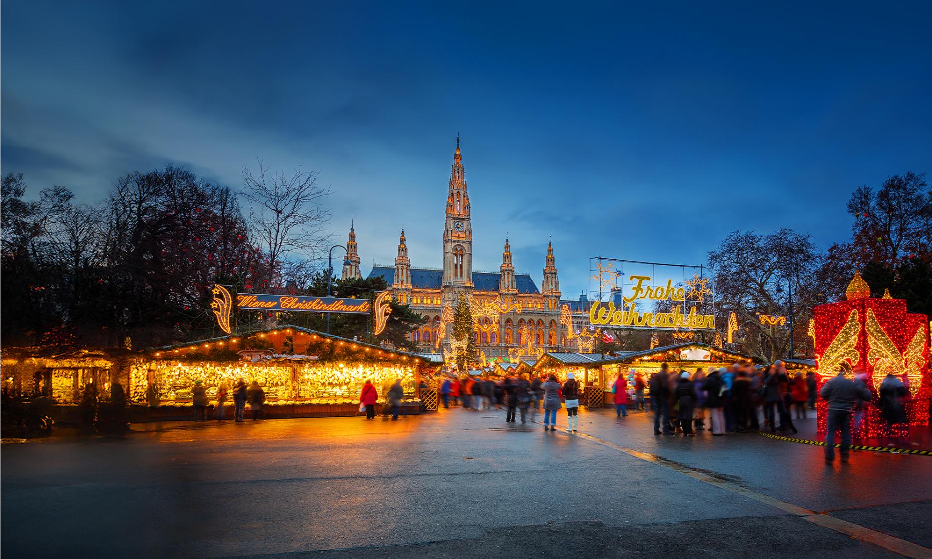 Christmas market in front of Vienna’s City Hall at dusk, with festive lights, decorated stalls, and crowds enjoying the festive atmosphere. Signs read “Wiener Christkindlmarkt” and “Frohe Weihnachten"