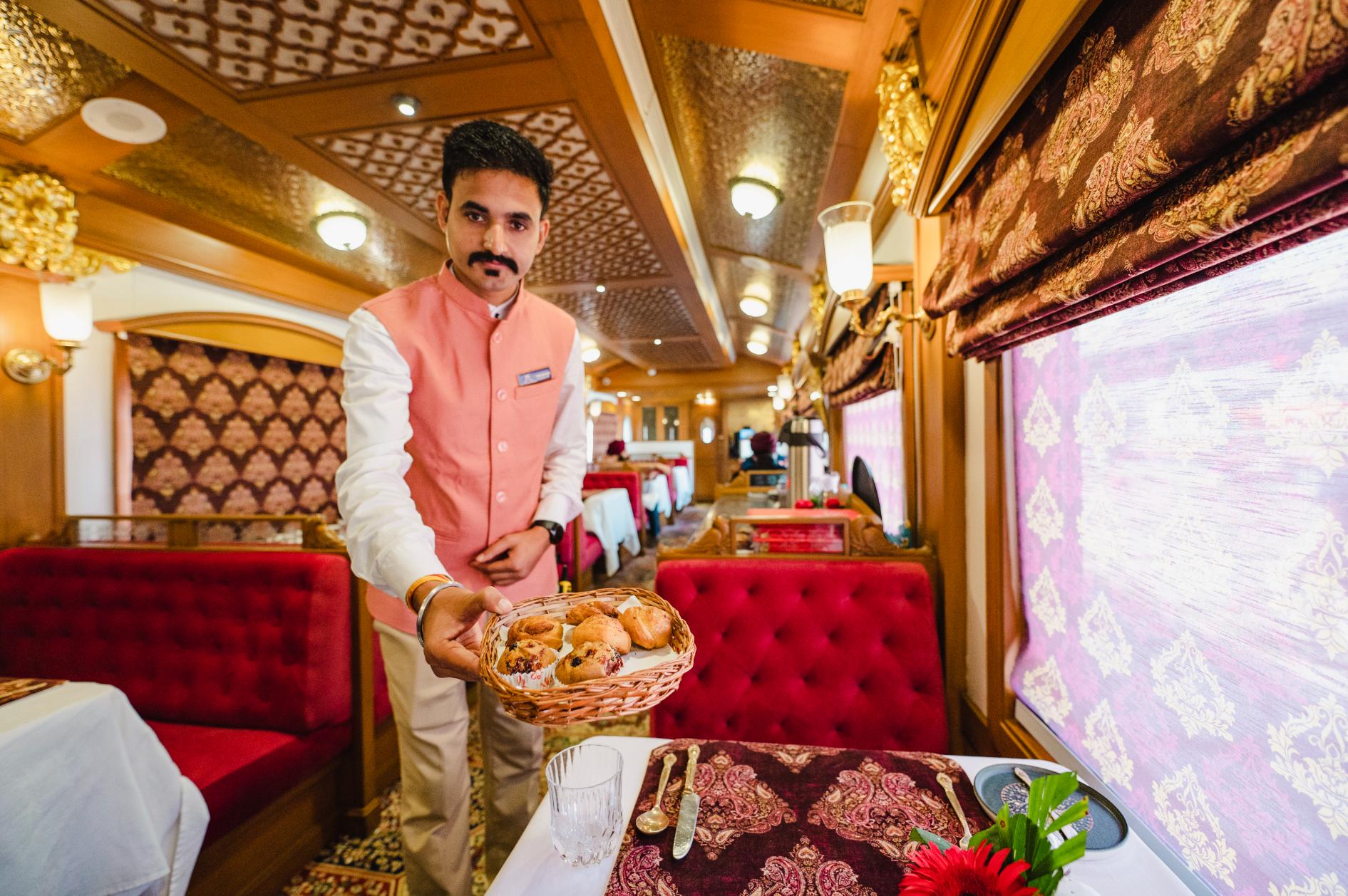 Dining aboard the elegant Deccan Odyssey train, with a server offering a basket of pastries. The dining car features red upholstered seats, red tablecloths, and matching decorative details