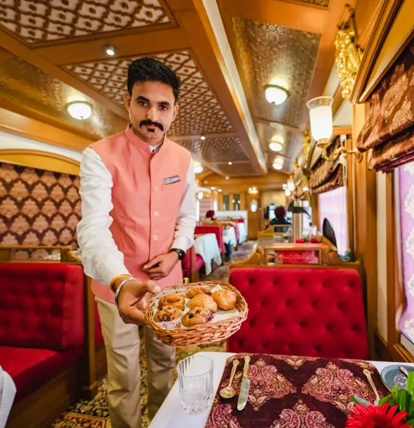 Dining aboard the elegant Deccan Odyssey train, with a server offering a basket of pastries. The dining car features red upholstered seats, red tablecloths, and matching decorative details