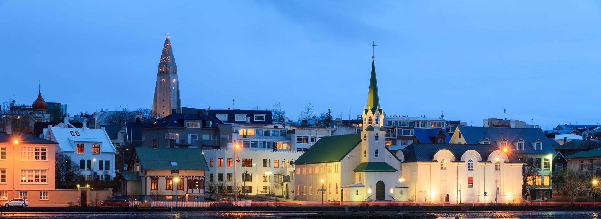 View of a town with a white church with a green roof at the front and a tower to the left sticking out above other buildings. Light blue evening sky