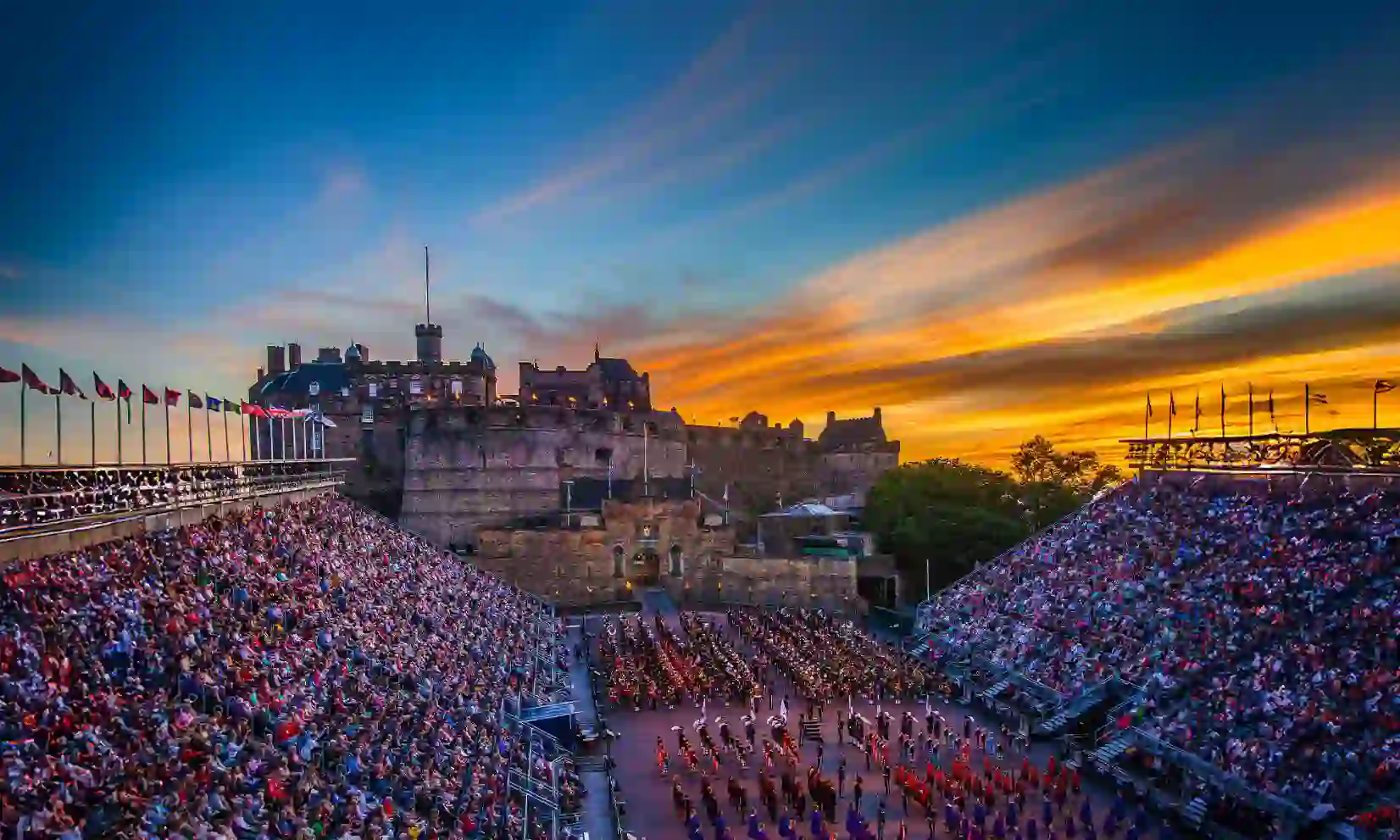 Edinburgh Castle at sunset overlooking a military parade during the The Royal Edinburgh Military Tattoo, with packed grandstands and rows of performers in uniform