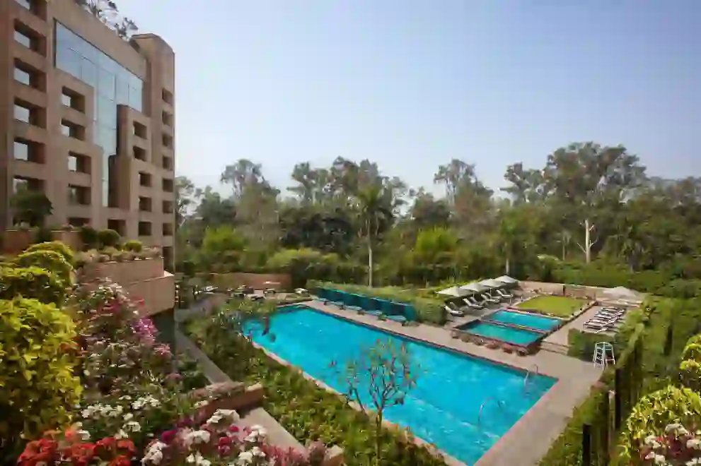 A view of the swimming pool and exterior of the Sheraton New Delhi, surrounded by landscaped gardens on a bright day