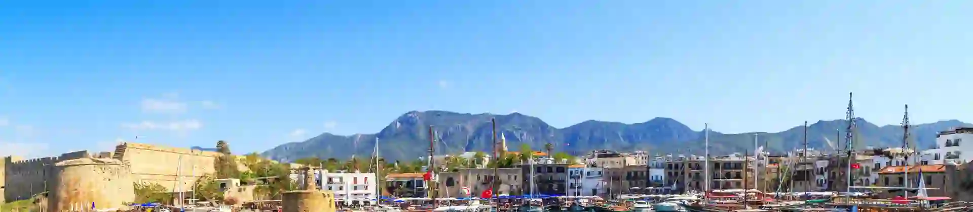 Boats docked at a harbour with bright blue water, buildings on the other side, and ruins of a castle to the left. Mountains in the distance in front of a blue sky.