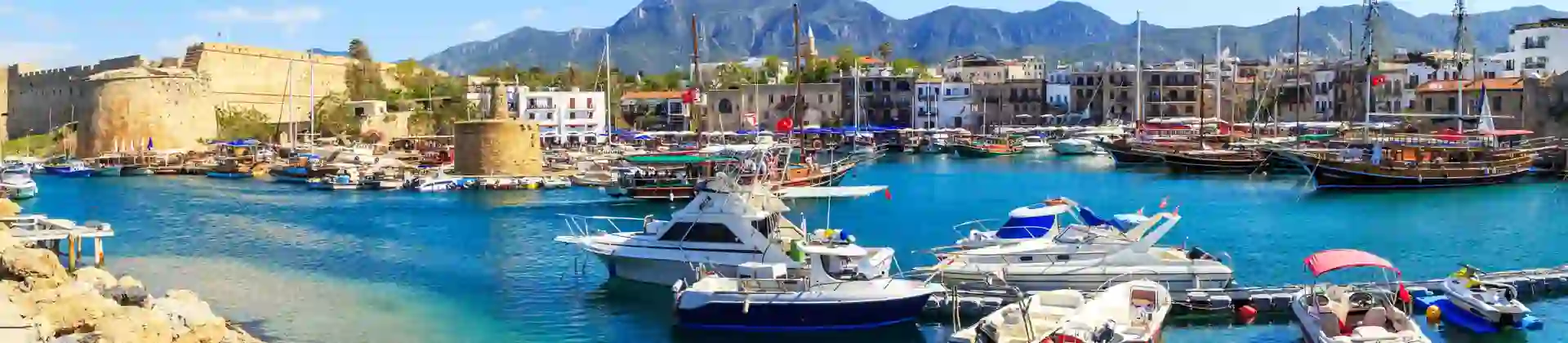 Boats docked at a harbour with bright blue water, buildings on the other side, and ruins of a castle to the left. Mountains in the distance in front of a blue sky.