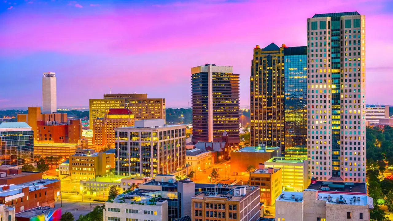 A view of the Birmingham, Alabama skyline at dusk, with skyscrapers and high-rises glowing beneath a vibrant pink and blue sky