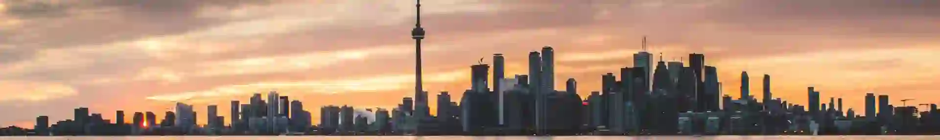 View over the water towards the Toronto skyline at sunset, with the CN Tower and surrounding buildings silhouetted against a pink and orange sky