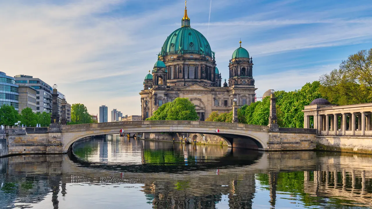 View of Berlin Cathedral, which is grey with turquoise domed roofs, and has a gold turret in the middle. In front is a bridge, and there is water in the forefront