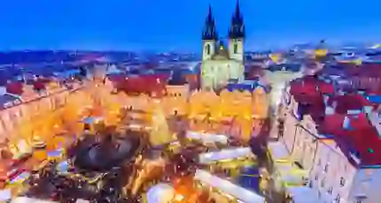 High angle shot of a Christmas market in the town square of Prague, showing a lit up gothic church with two towers and a large lit up Christmas tree