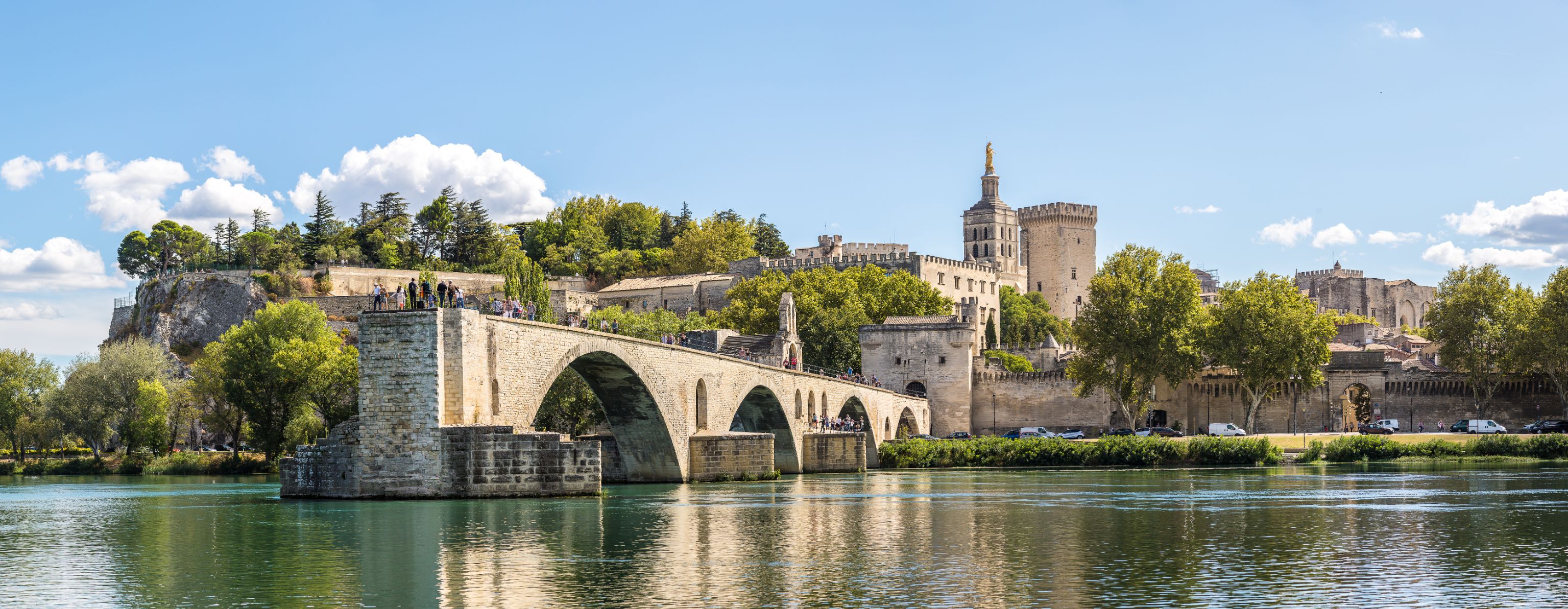 Wide angle shot of the Pont d'Avignon bridge, with the palace on the other end. The river water in the forefront, and a blue sky with clouds above.