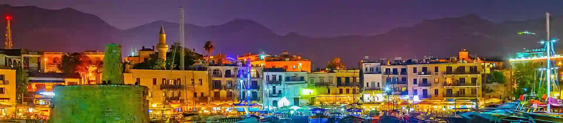 View of a harbour at night, with boats docked and a ruin to the left. Multi coloured lights behind the boats, reflecting on the water. A strip of buildings on the far side of the harbour, in front of a silhouette of mountains in front of a night sky.