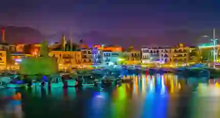 View of a harbour at night, with boats docked and a ruin to the left. Multi coloured lights behind the boats, reflecting on the water. A strip of buildings on the far side of the harbour, in front of a silhouette of mountains in front of a night sky.