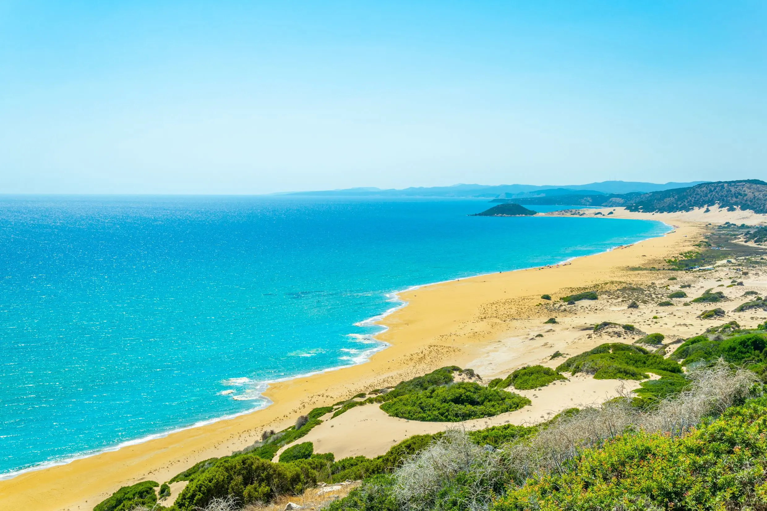 High angle shot of a strip of beach, with yellow sand and bright blue water, leading towards mountains in the distance. The land behind the beach the right forefront has patches of grass and sand.