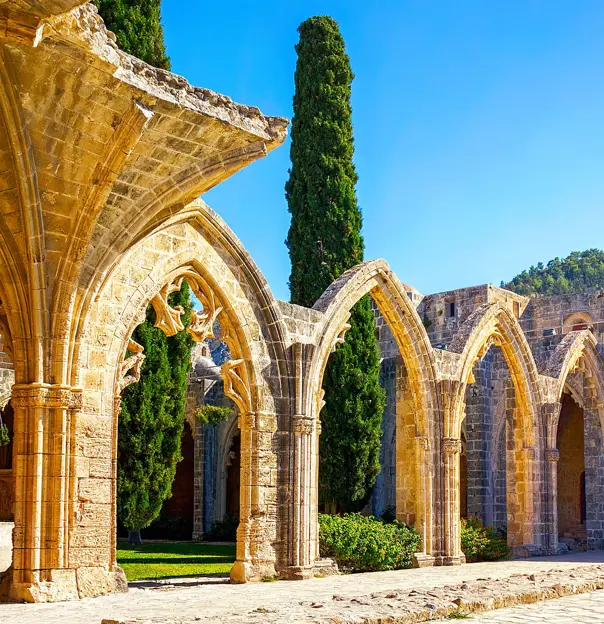 Low angle shot of the ruins of four brick archways connected to a wall with steps attached to it, and a thin, rectangular structure on top. Behind is the view of the mountain in front of a blue sky. To the left, behind the archway, two tall dark green trees.