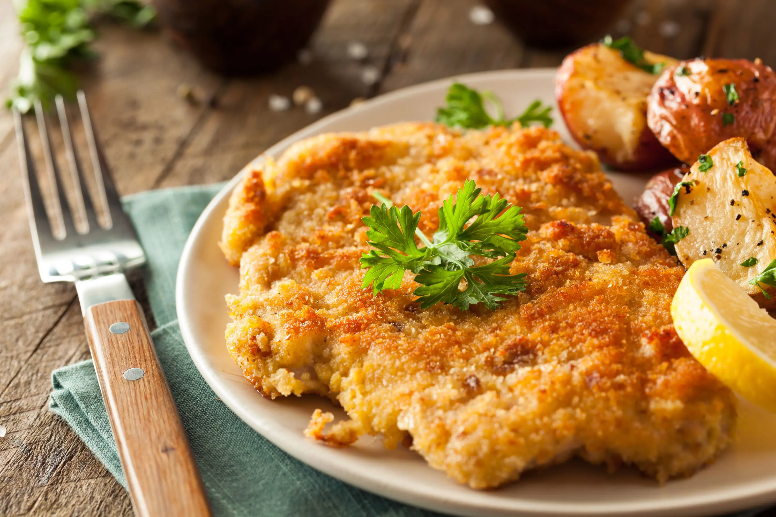 Close up of half a plate of food, with a shnitzel and a lemon and some potatoes next to it. A fork to the left of the dish with a turquoise napkin underneath it.
