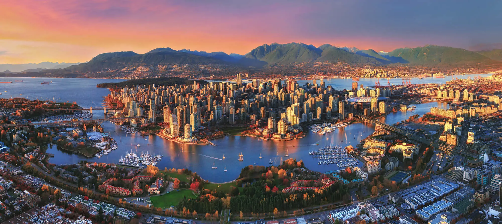 Aerial view of Vancouver, Canada at sunset, showing the city skyline, surrounding water, and distant mountains beneath a glowing, colourful sky