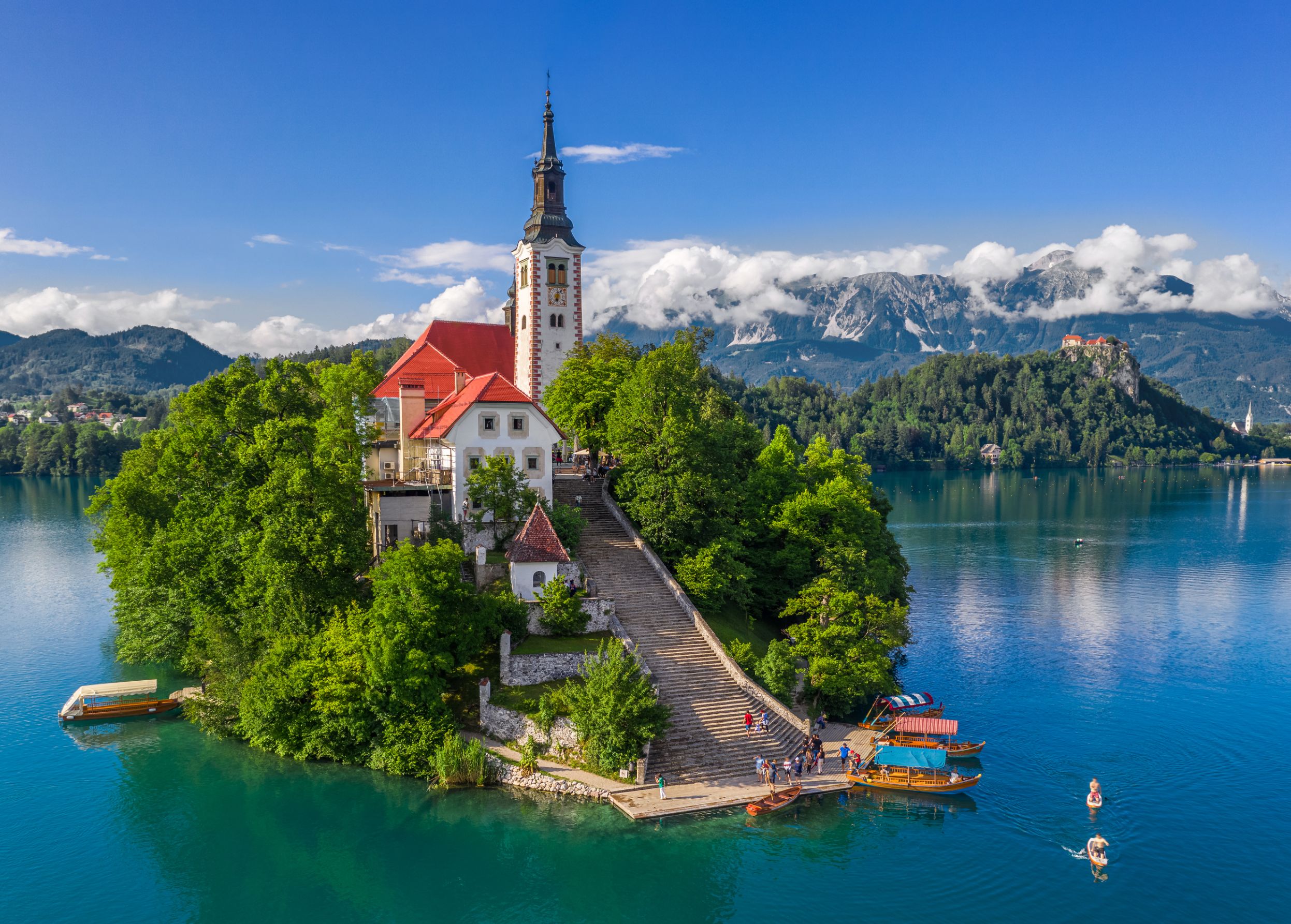 Shot of a church on a small bit of forested land on water, there is a pathway leading up to the church which has some people and boats at the bottom. Behind, there are mountains and clouds, under a blue sky. 
