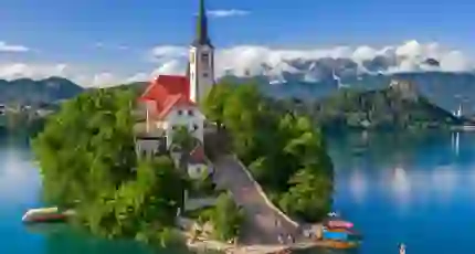 Shot of a church on a small bit of forested land on water, there is a pathway leading up to the church which has some people and boats at the bottom. Behind, there are mountains and clouds, under a blue sky.