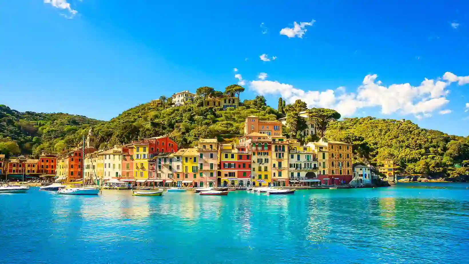 A vibrant coastal village in Portofino, Italy, with colourful buildings lining a harbour filled with small boats, set against a lush green hillside under a bright blue sky