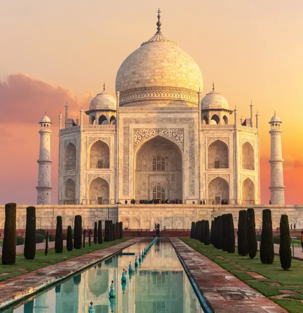 The Taj Mahal in Agra glowing in the warm hues of sunset, its white marble reflecting the golden light, with the surrounding minarets and the long reflecting pool in front adding to its majestic symmetry
